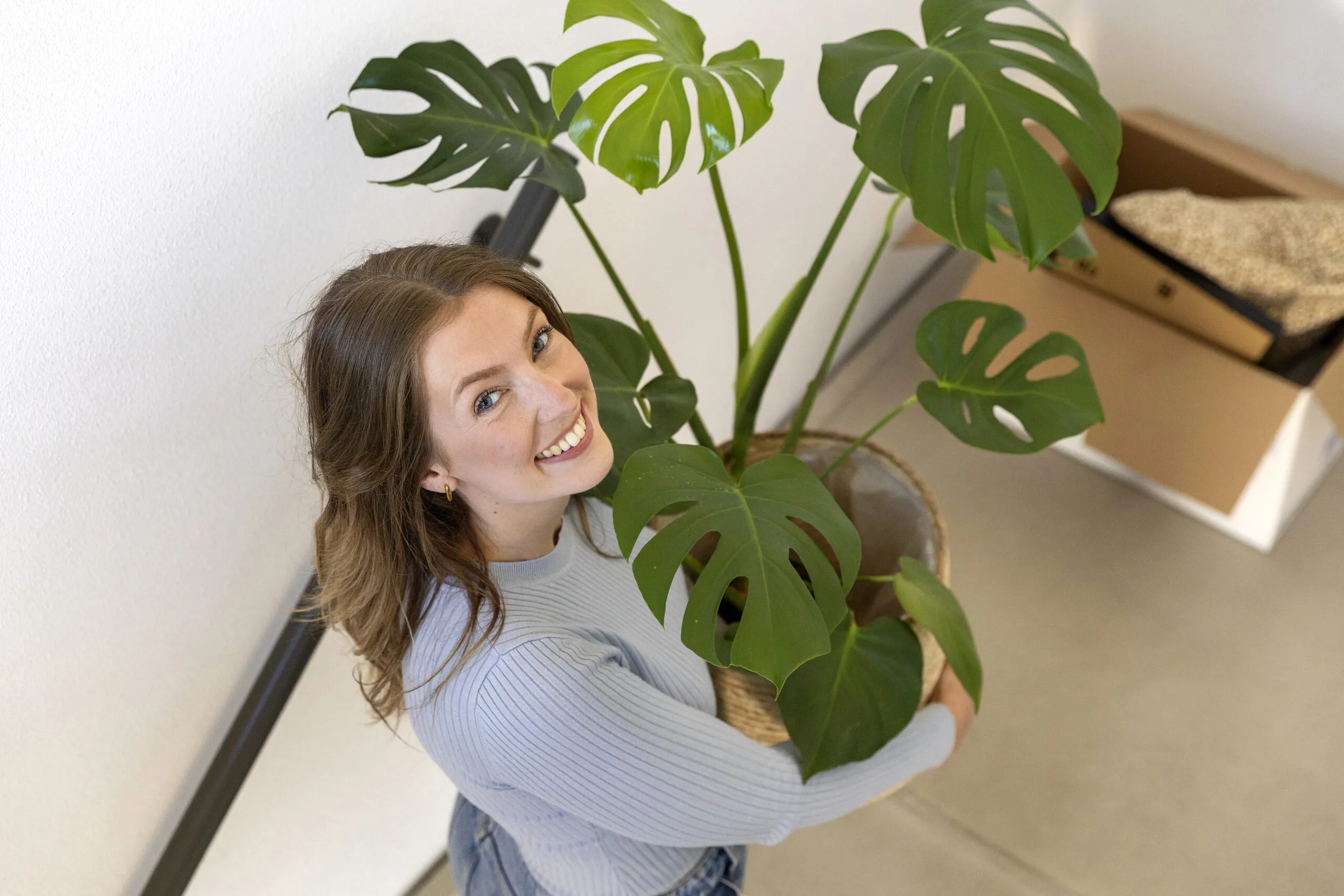 Vrouw lacht naar de camera met een grote plant in haar handen.