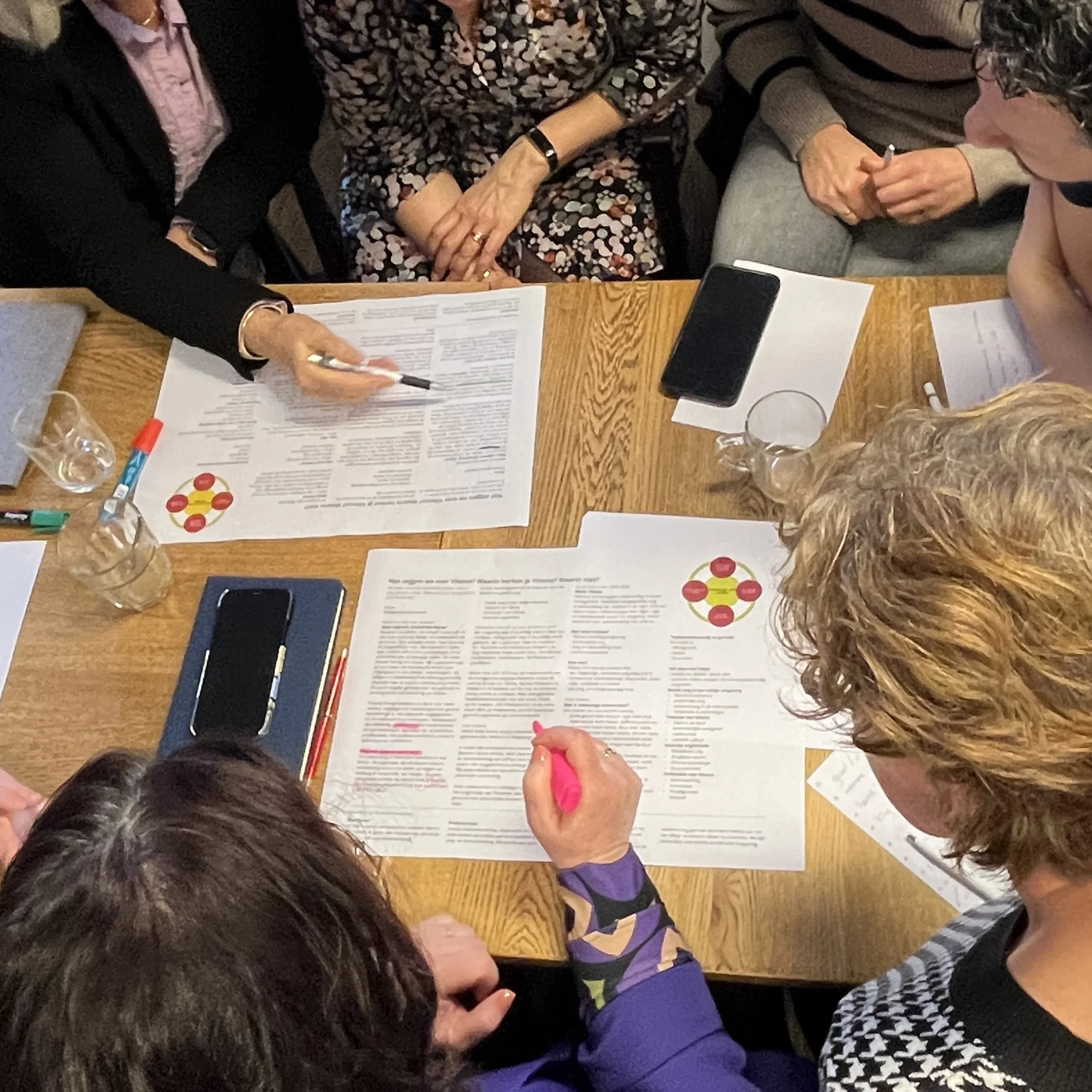 Meerdere mensen gebogen over vellen papier op tafel, met pennen en highlighters in de hand.