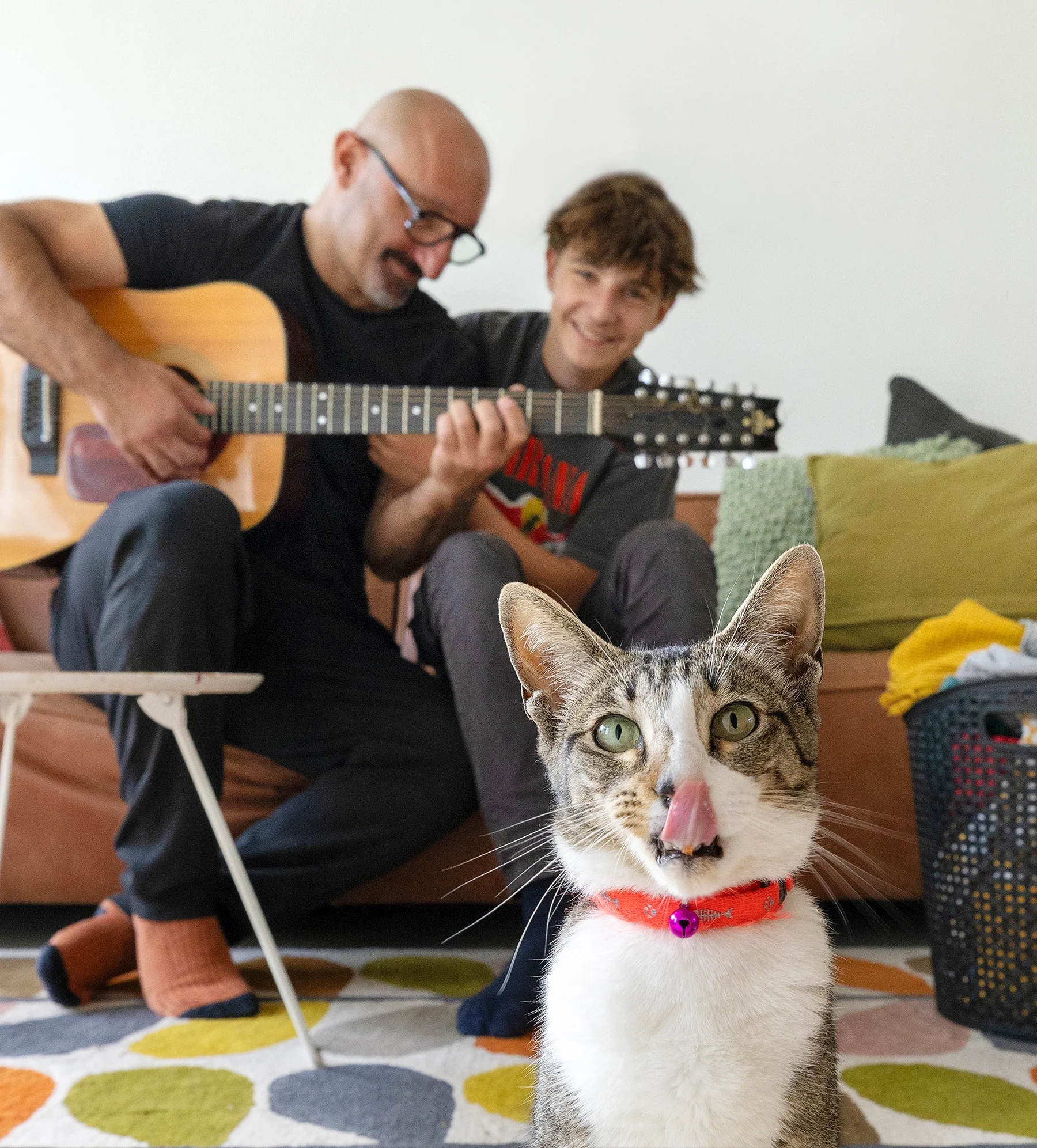 Vader en zoon zitten op de bank en spelen gitaar- op de voorgrond een kat die de camera inkijkt