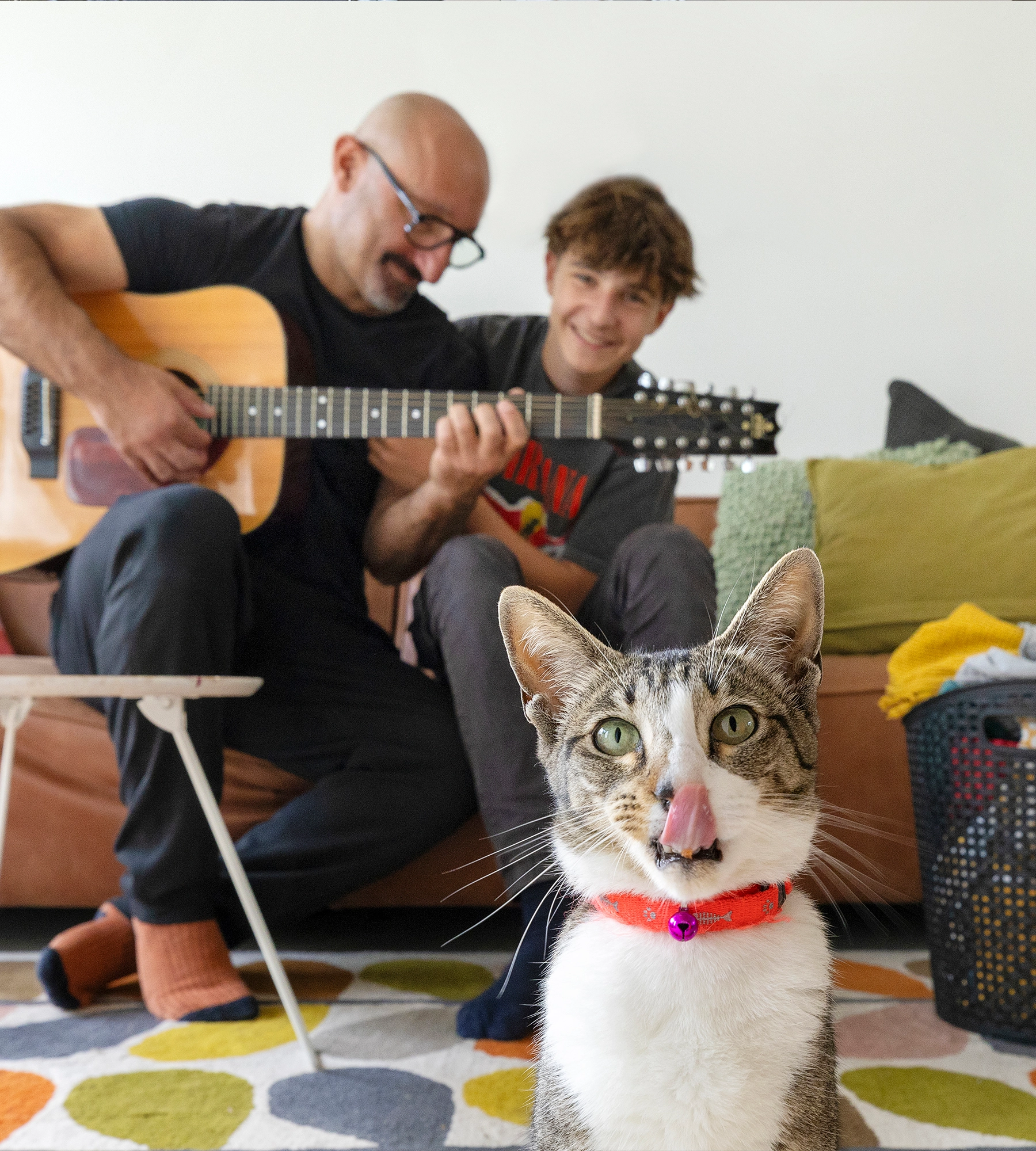 Vader en zoon zitten op de bank en spelen gitaar- op de voorgrond een kat die de camera inkijkt