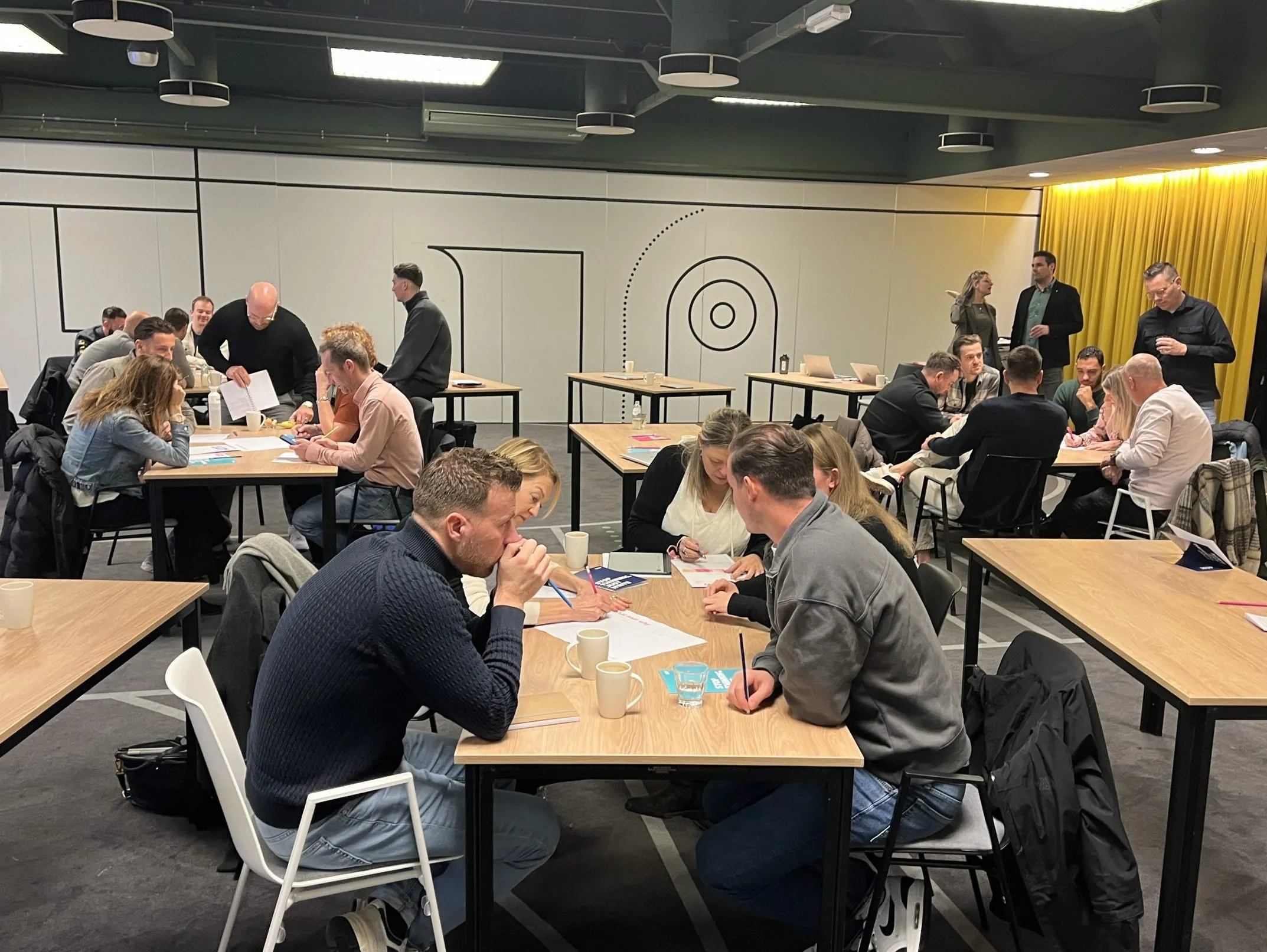 Deelnemers overleggen in kleine groepjes aan tafels tijdens een werksessie in een moderne vergaderruimte.