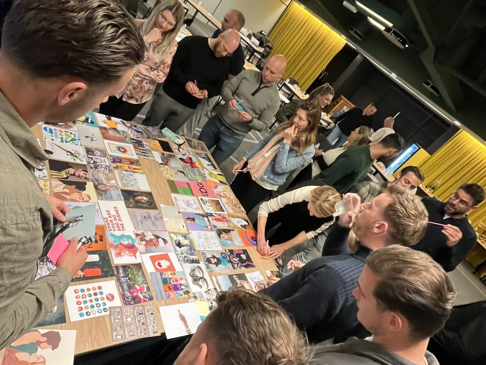 Deelnemers staan rond een tafel met afbeeldingen en bespreken hun keuzes tijdens een creatieve werksessie.